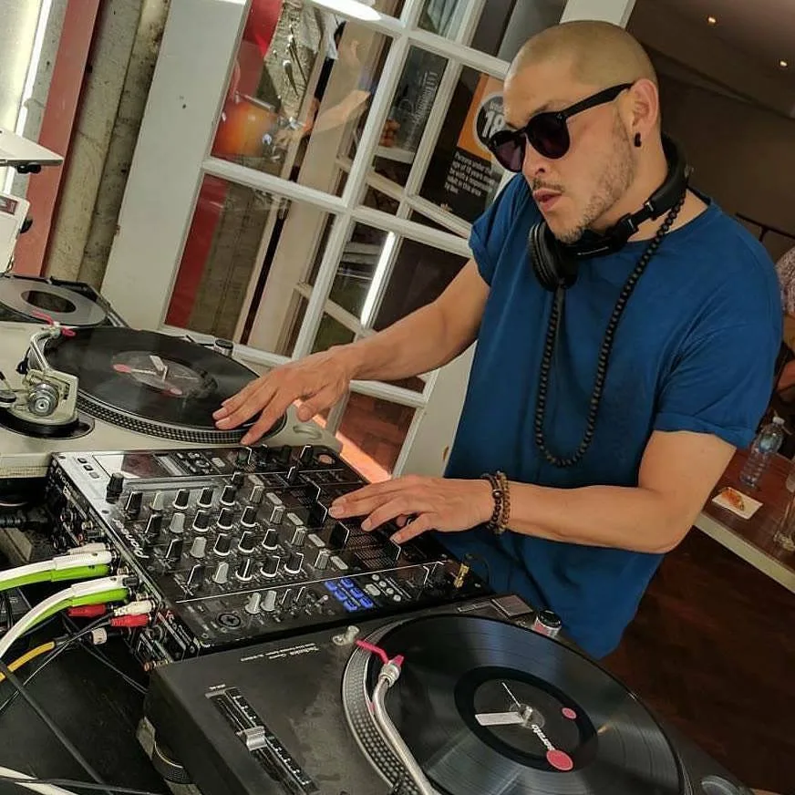 DJ CMAN portrait behind the decks in a blue shirt, sunglasses round his neck