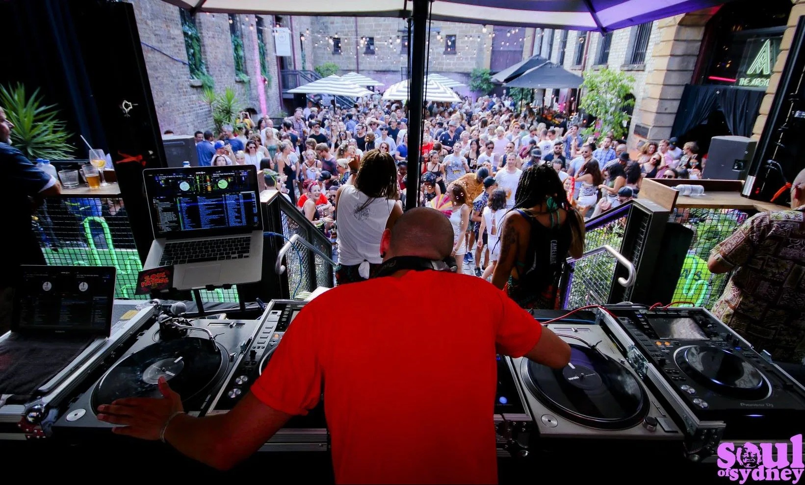 DJ CMAN DJing to a packed courtyard crowd, string lights strung overhead