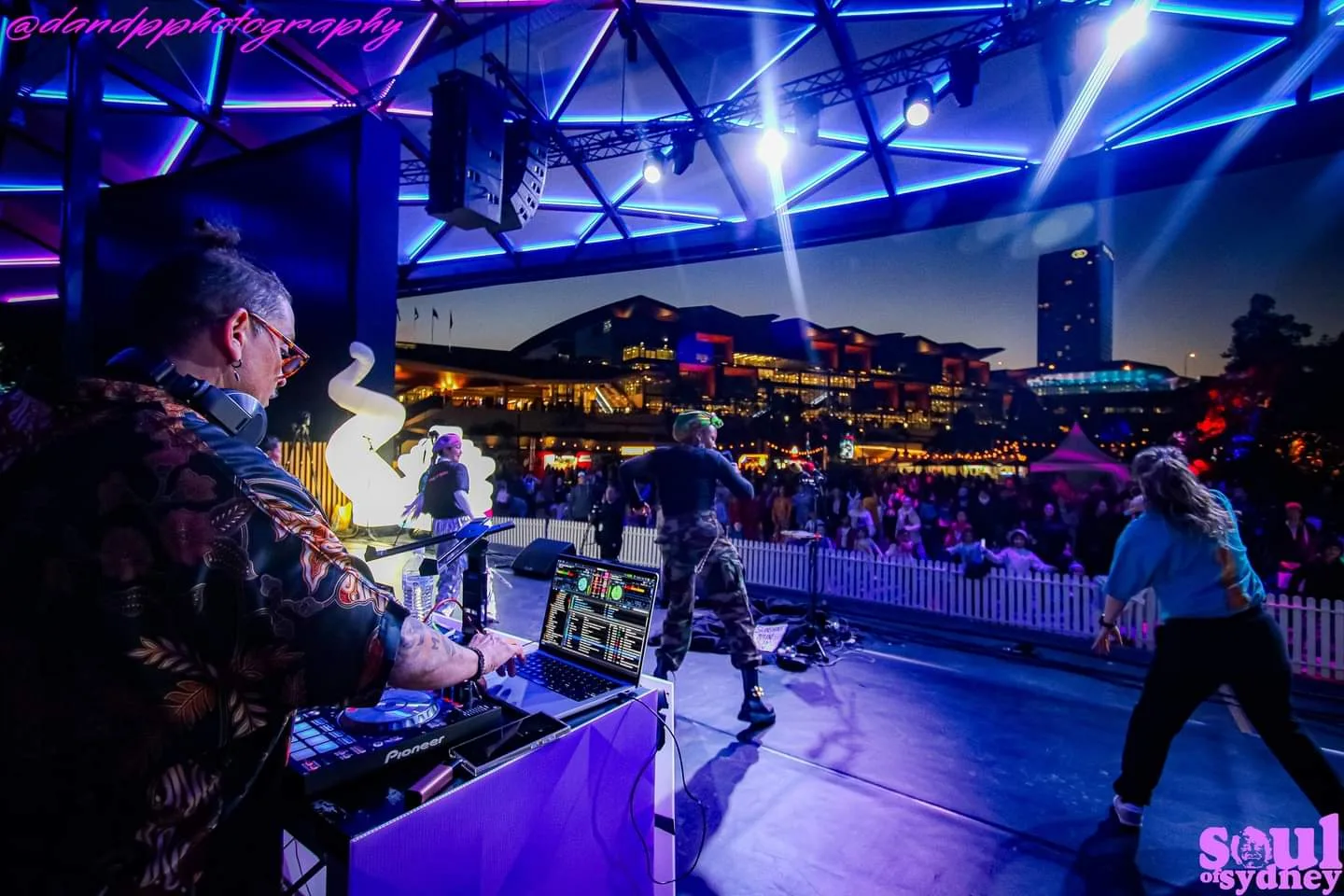 DJ CMAN DJing an evening outdoor stage set, dancers beside him and the lit-up city skyline behind the crowd