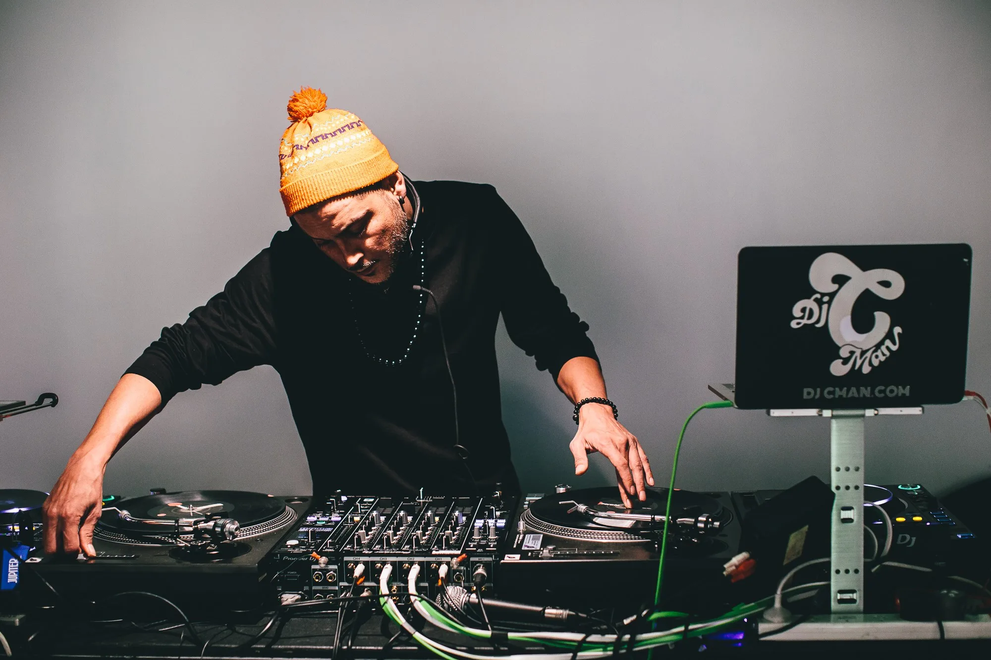DJ CMAN spinning vinyl on twin turntables in a studio portrait, wearing an orange beanie