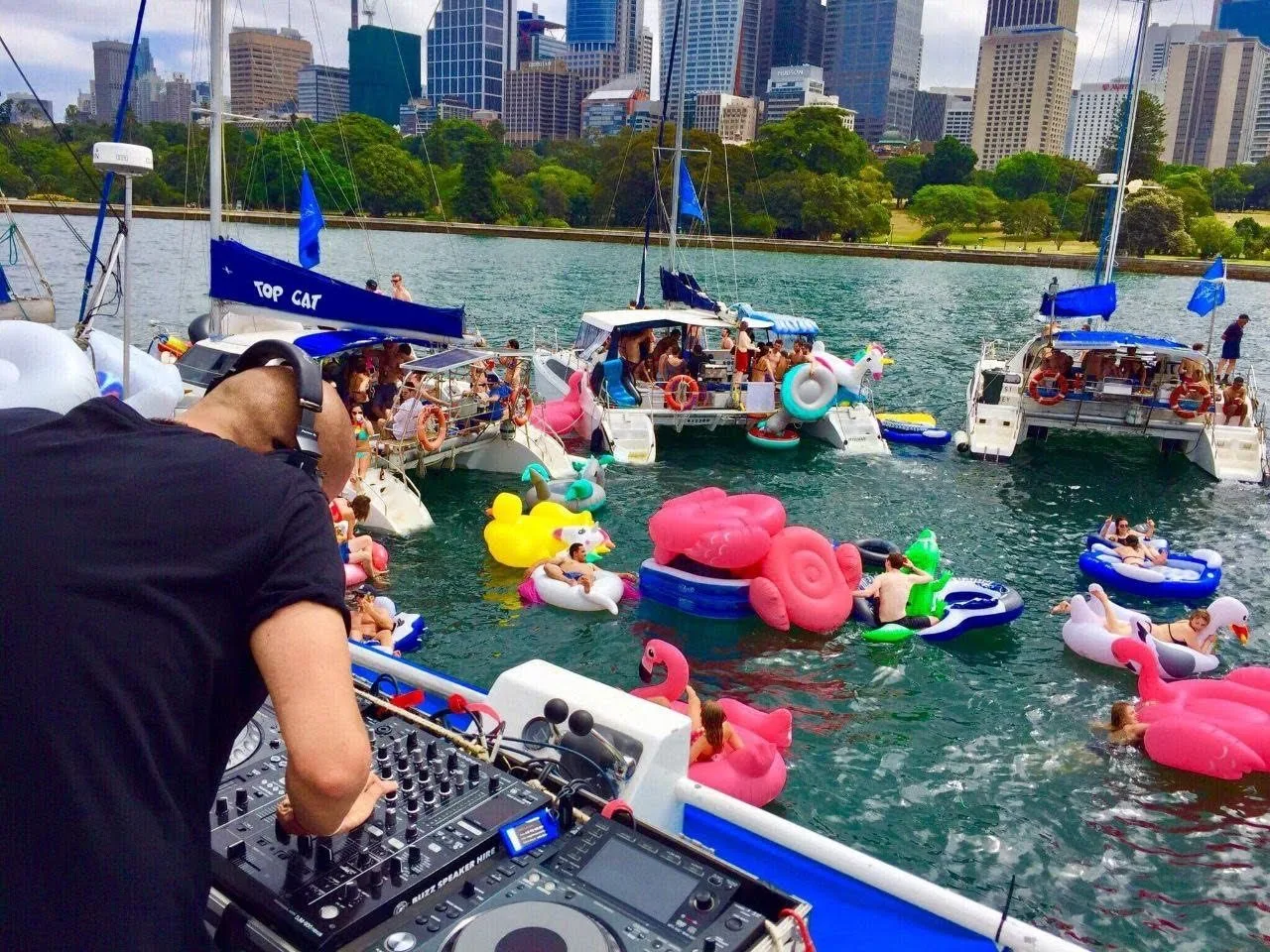 DJ CMAN DJing a Yacht Club Social boat party on Sydney Harbour, catamarans and inflatables rafted up with the city skyline behind