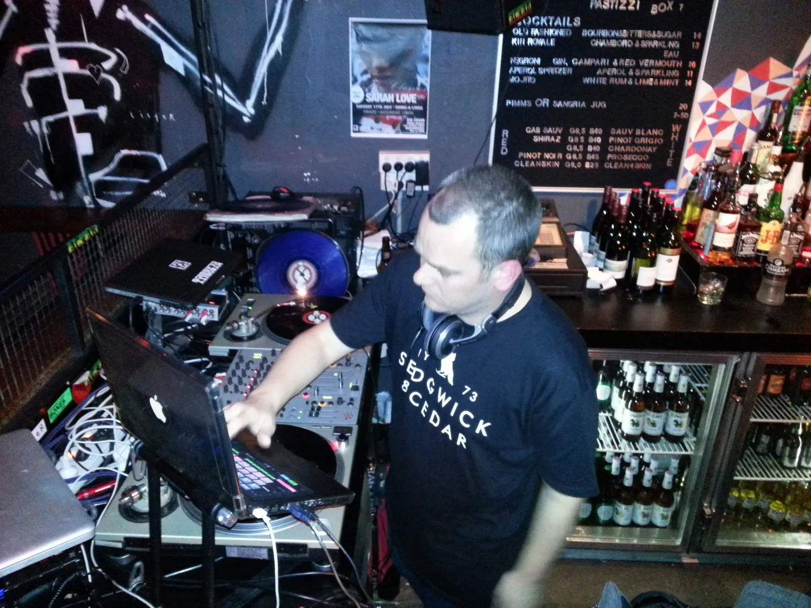 A DJ leaning over the laptop and mixer in the Ching-a-Lings booth, bottles racked behind the bar
