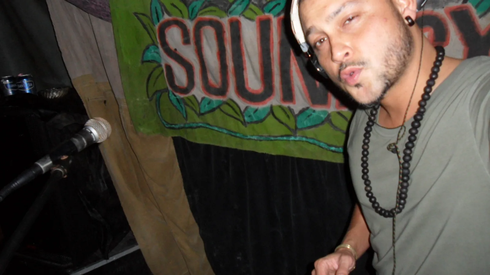 DJ CMAN with headphones on beside a microphone, a painted sound system banner behind him