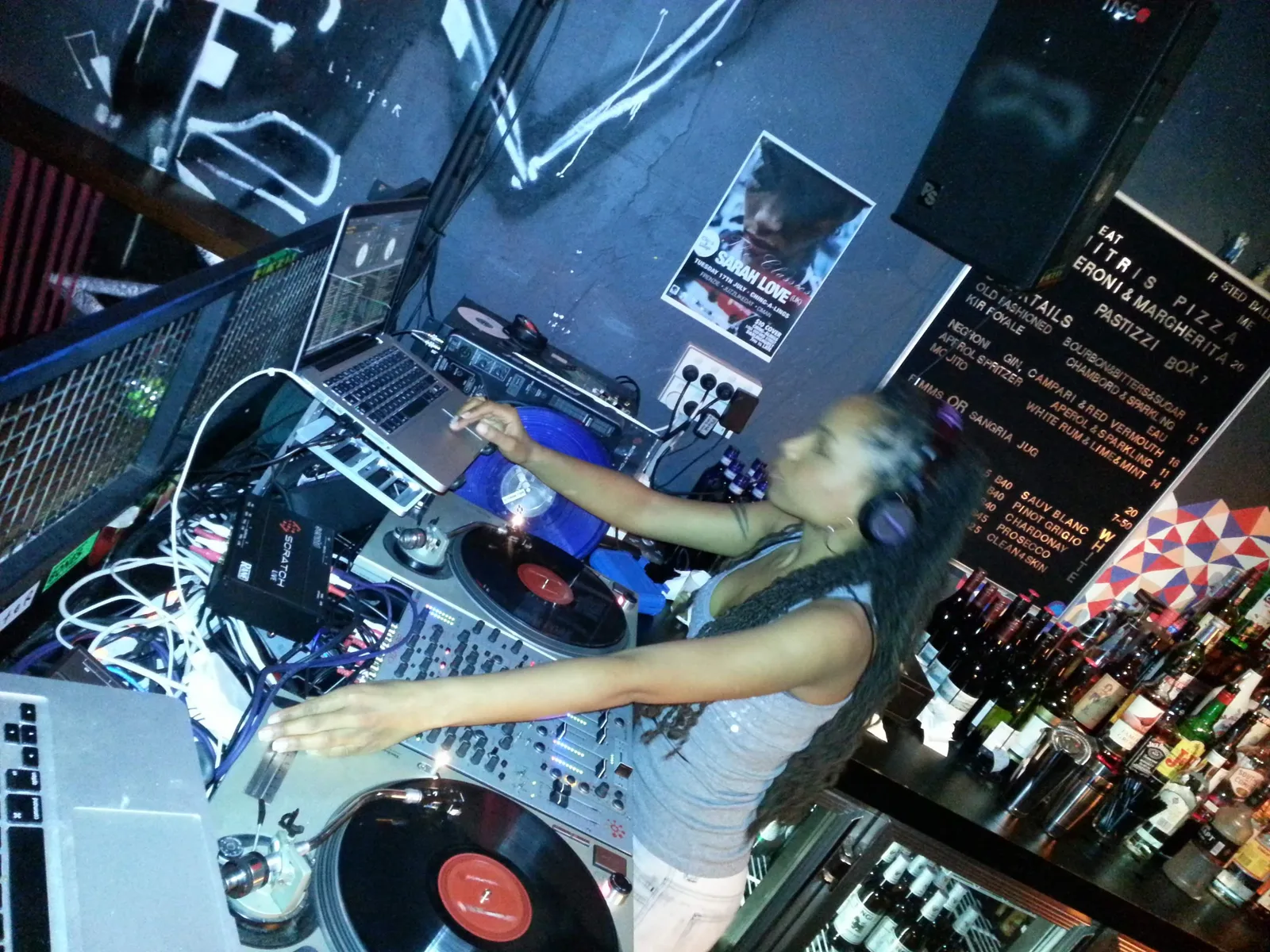 A DJ mid-set behind the decks at Ching-a-Lings, records and mixer lit up under the bar's cocktail board