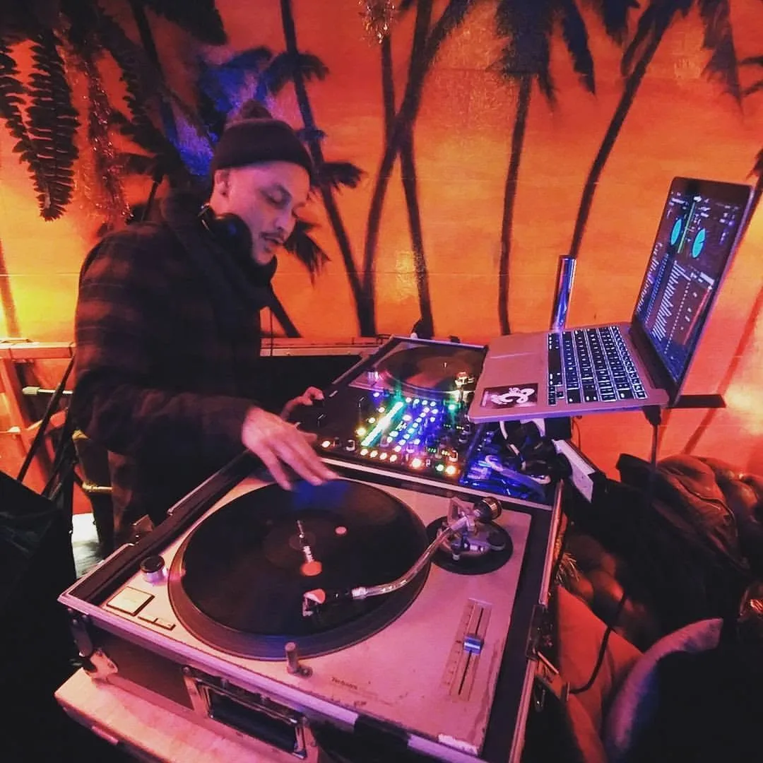 DJ CMAN alone at the decks against Cafe Lounge's orange sunset palm mural.
