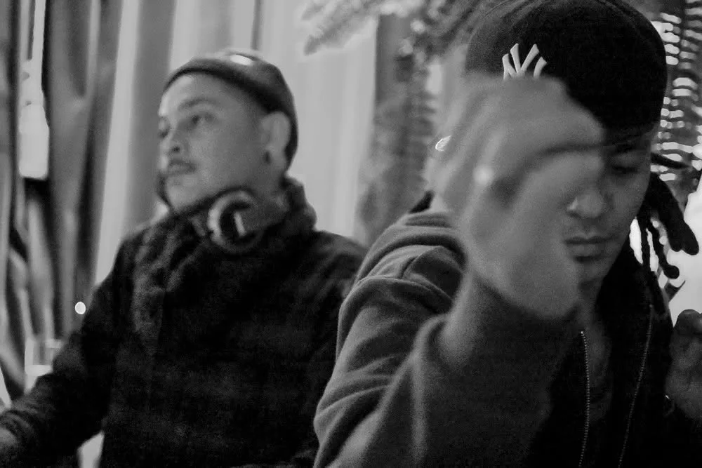 Black and white moment backstage, JuzzLikeDat blurred mid-gesture with DJ CMAN seated behind.