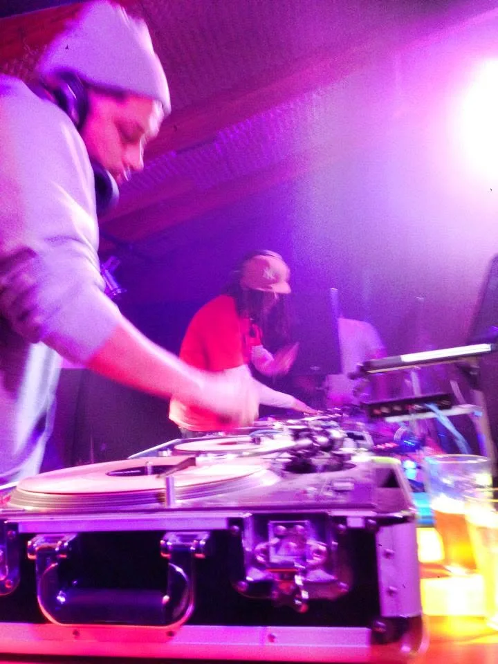 Close on DJ CMAN cueing a record, JuzzLikeDat watching from behind the booth.