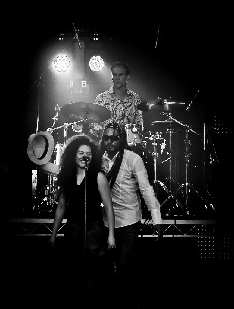 Cumbiamuffin performing live, two vocalists at the microphones with the drummer lit behind them, black and white
