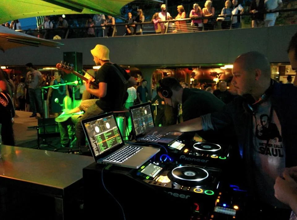 DJ CMAN at the decks in the Opera Bar booth at night, with the crowd on the balcony above