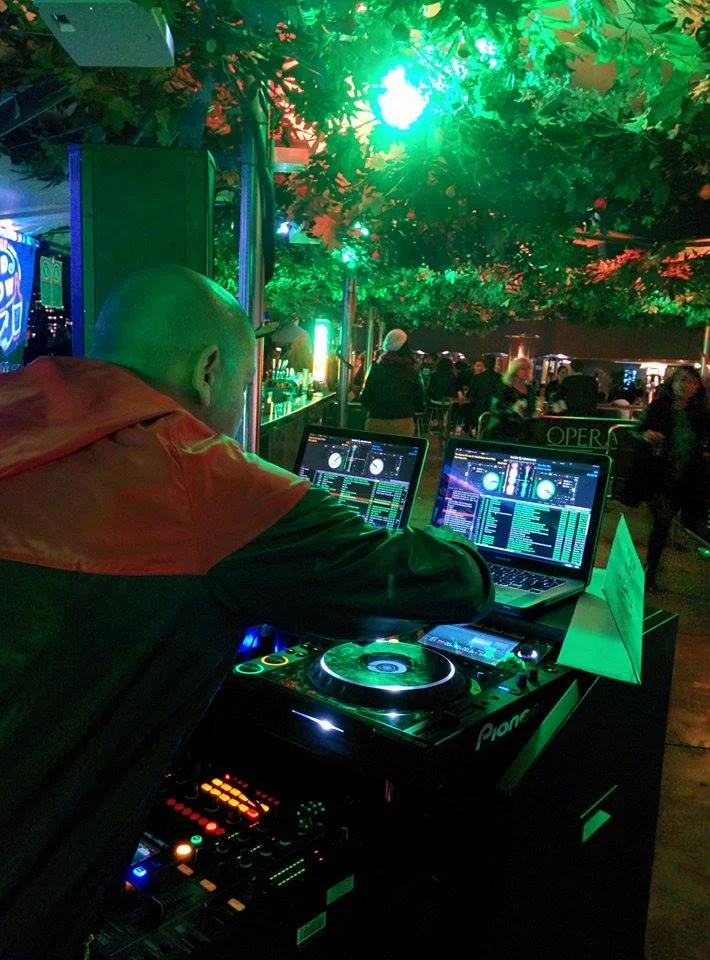 Photo of CMAN DJing at Opera Bar under the vine canopy at night, leaning over two laptops and a Pioneer CDJ lit green, with the harbourside bar crowd behind