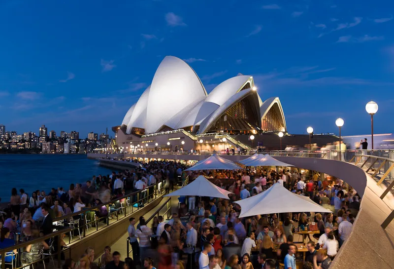 Crowd on the terrace at Opera Bar at dusk, with the Sydney Opera House sails and the city skyline across the water