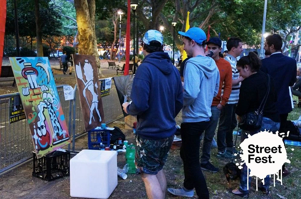A crowd gathered at dusk beside live-painted canvases propped along a fence at Street Fest