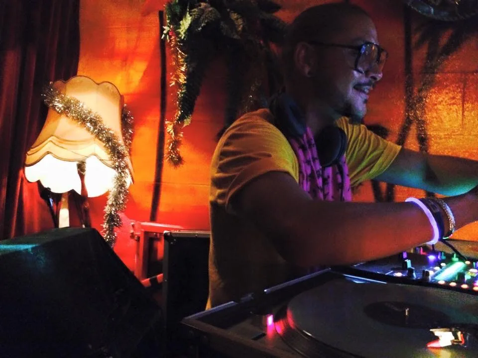 DJ CMAN at the decks in a yellow tee and lei, playing in front of Cafe Lounge's painted palm mural
