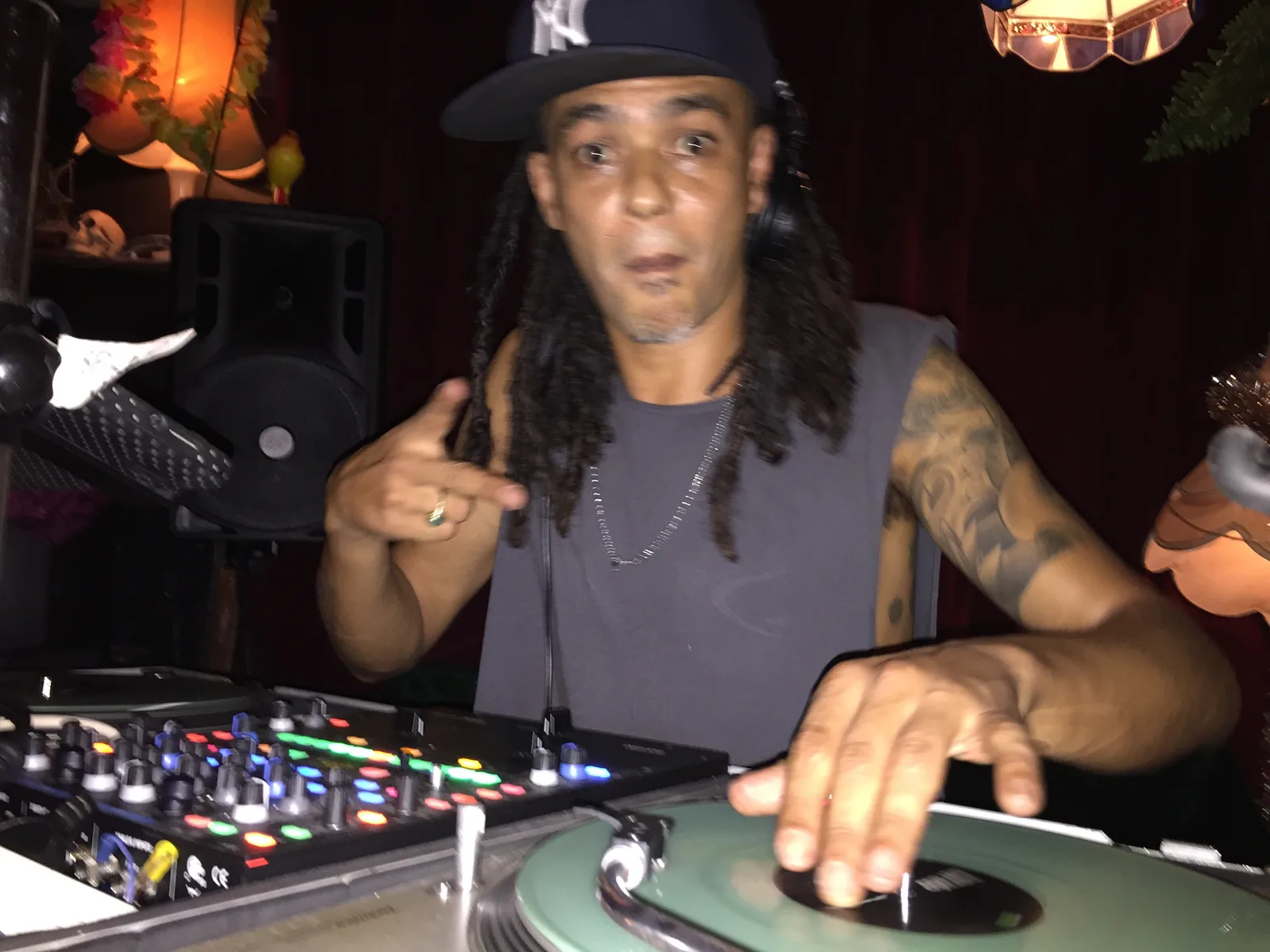 JuzzLikeDat at the turntables in a cap and singlet, one hand on the record, pointing at the camera