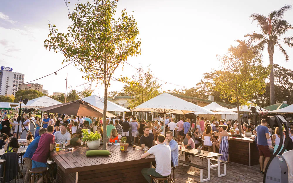 Crowd at long communal tables in the beer garden at The Greens North Sydney at sunset, with marquees and festoon lights