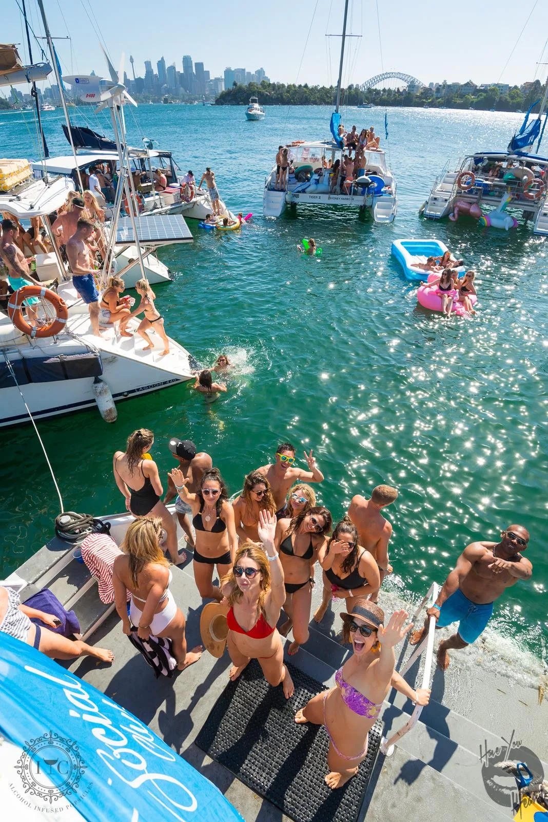 Crowd on the deck of a catamaran at The Yacht Social Club raft-up, with the Sydney Harbour Bridge and city skyline behind