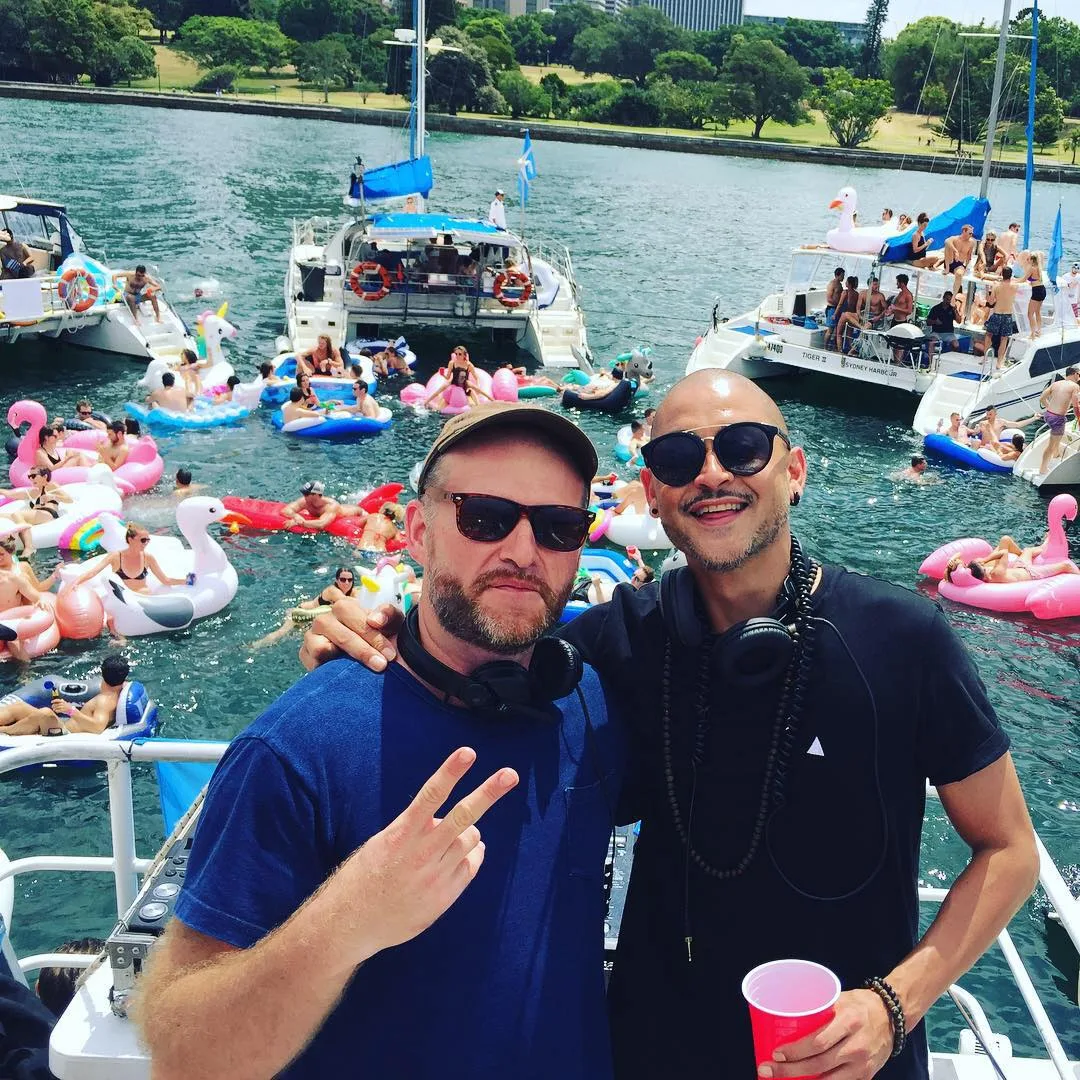 Two DJs on deck with headphones, one throwing a peace sign