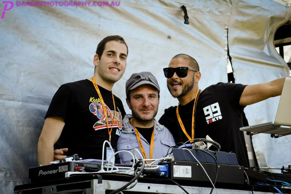 Phil Toke in a flat cap posed with two friends crowded into the DJ booth behind the Pioneer decks