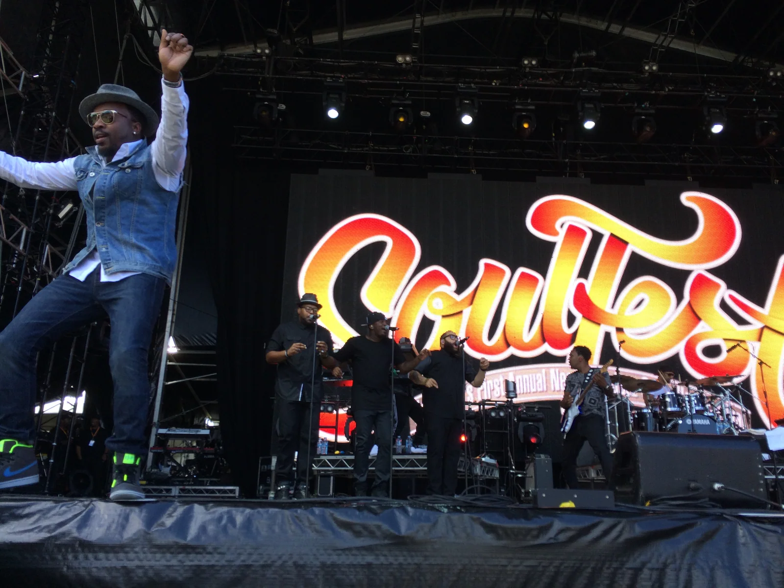 Anthony Hamilton in a hat and denim vest leaping at the front of the Soulfest stage, his band and the Soulfest logo screen behind