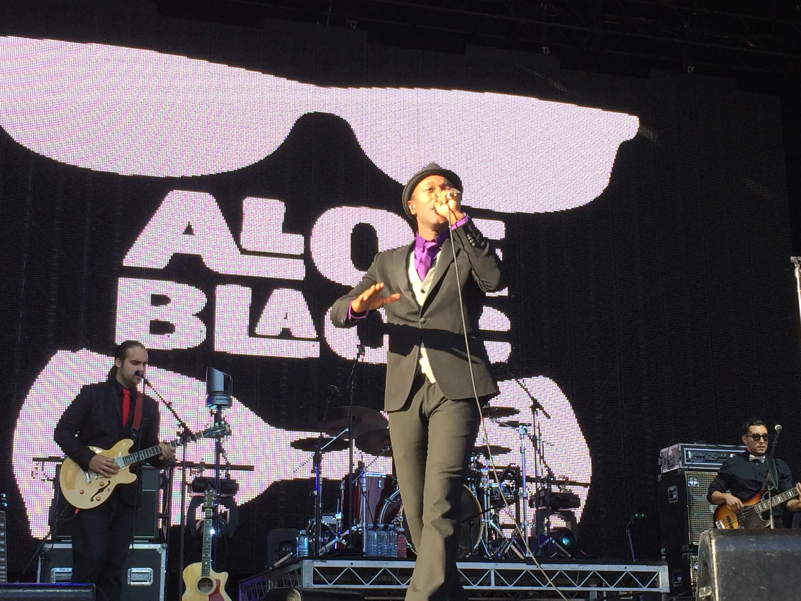 Aloe Blacc singing in a grey suit and hat in front of a giant Aloe Blacc screen with his band around him