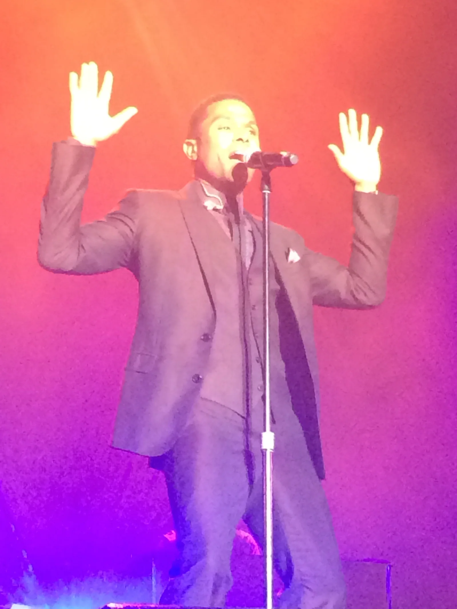 Maxwell at the mic stand with both hands raised, washed in red and purple stage light